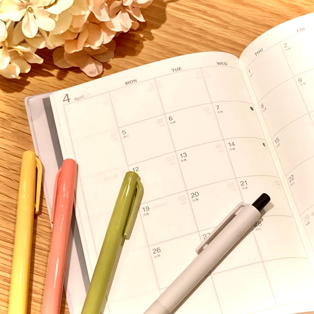 【テラスモール湘南】春の日に手帳を書き込むところをイメージした、白とピンクの花、商品のマンスリーウィークリースケジュール帳、水性サインペンのうぐいすいろ、あんずいろ、からしいろ、黒のボールペンがテーブルに置かれた写真