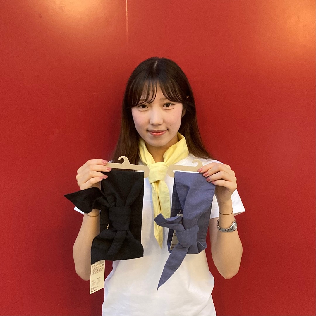 黄色のスカーフを首に巻いて、黒と青のスカーフを手に持っている女性スタッフ
