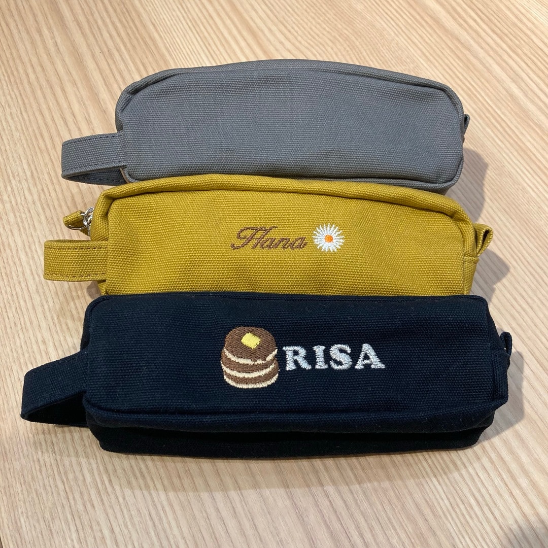 【テラスモール湘南】帆布ペンケースの、刺繍を施していないグレー、名前と花のマークを入れたマスタードイエロー、ホットケーキのマークと名前を入れたネイビーを縦に並べた写真