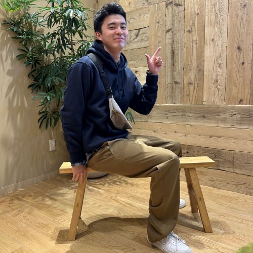 ネイビーのトレーナーにブラウンのパンツをはいて椅子に座る男性