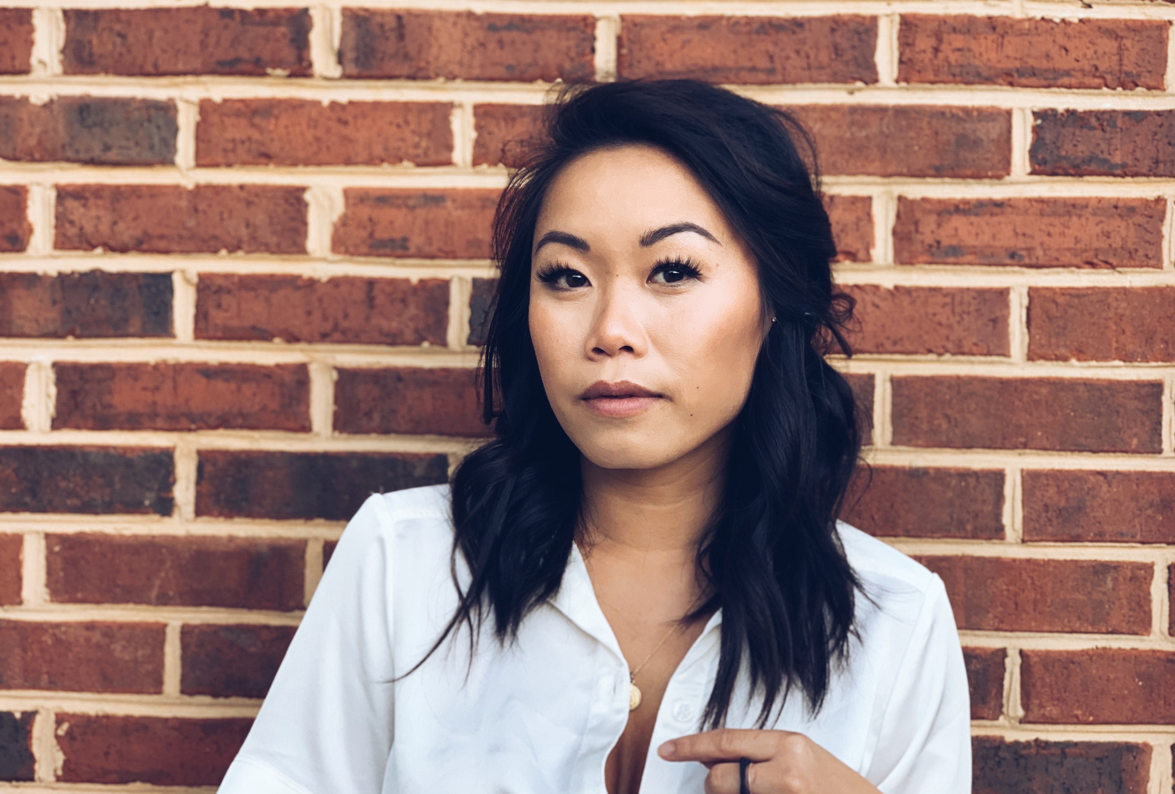 Asian woman stands in front of an exposed brick wall. Her hair is shoulder length, wavy, and black. She has on a white v-neck tshirt.