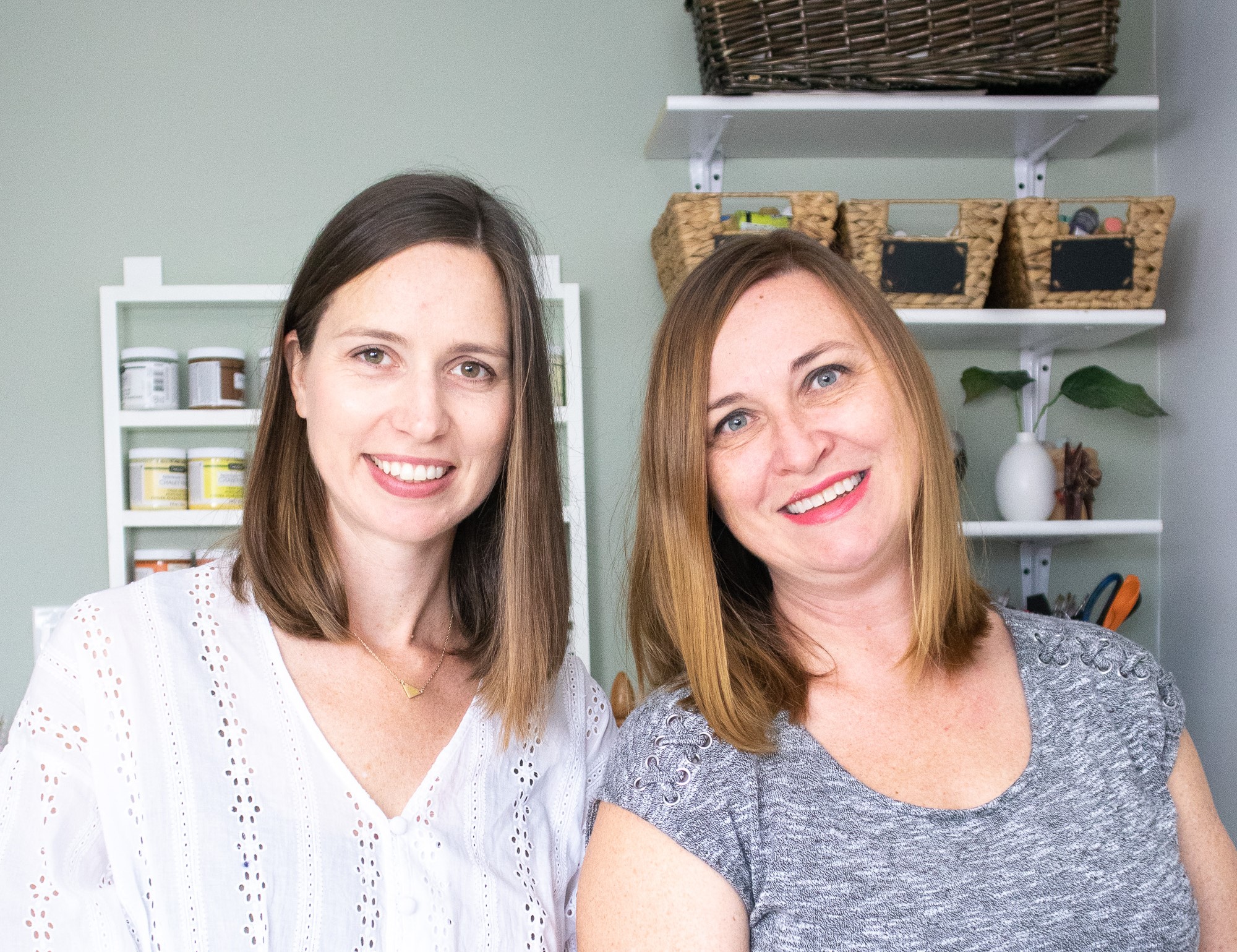 Jane and Sonja stand side by side. Jane wears a white button up blouse and Sonja is wearing a grey crew neck t-shirt. They are standing in front of a grey wall with white mounted shelves. Crafting materials are on the shelves. Both women are facing the camera and smiling, with their heads slightly tilted toward each other.
